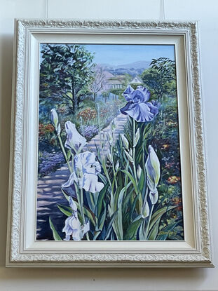 Purple Iris in the foreground with a path leading to Monet's house, lined with lush bedding plants and  trees, in soft lilacs, pinks and greens against a pale blue sky, on a portrait orientation canvas with a tradition carved cream coloured frame.