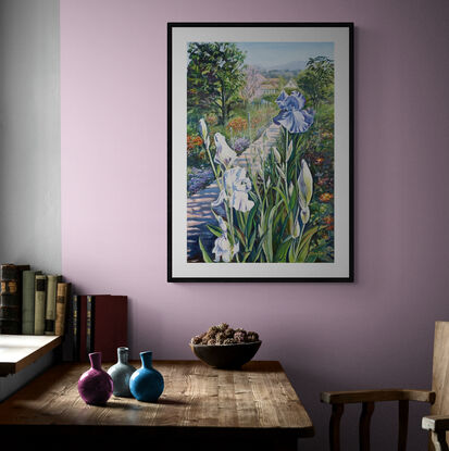 Purple Iris in the foreground with a path leading to Monet's house, lined with lush bedding plants and  trees, in soft lilacs, pinks and greens against a pale blue sky, on a portrait orientation canvas with a tradition carved cream coloured frame.