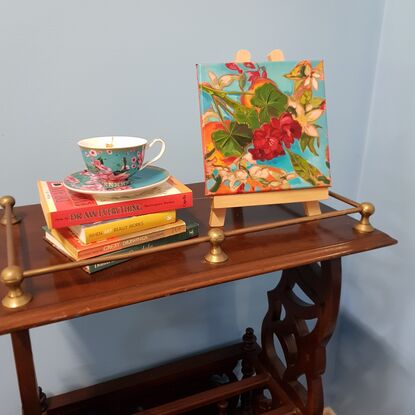 A still life arrangement with red geranium flowers with large dark green leaves, patterned aqua and orange scarf in the background.