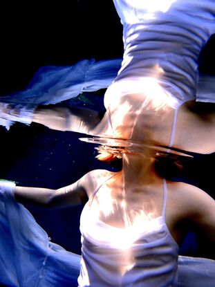 Young woman rising through the surface of the water in a white dress - capturing a mirrored reflection of light.