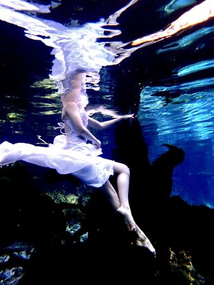 Young woman sitting on a log underwater in white dress whilst creating reflections in the surface.