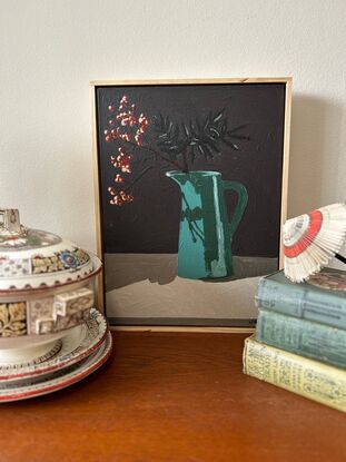 A tall deep aqua coloured vintage jug sits on a sideboard. In it are bare branches with berries light red in colour. They cast a shadow on the jug and the background is a dark grey.