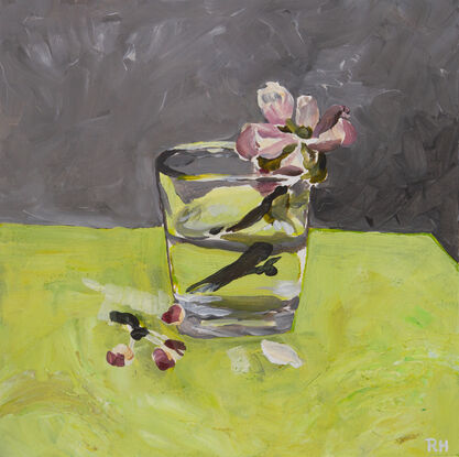 This is a single cherry blossom, in a glass of water, painted on a bench of bright green, with a grey backdrop.