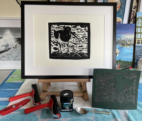 Lino Cut portrait of an Australian Pied Oystercatcher Bird standing on rocks with a background of swirling water. The bird has a very long beak. It’s feathers are black with a white breast and belly. 