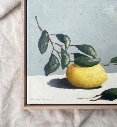 A lemon on a branch with leaves painted with subtle texture on a cream base and a blue grey background