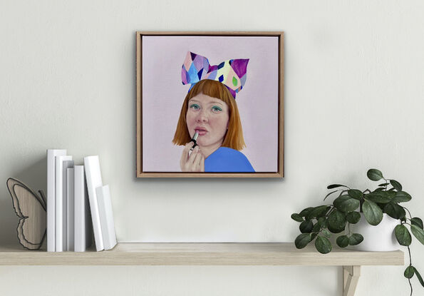 A beautiful red headed girl with bright green eyeshadow  and rosy cheeks is applying a peach colour lipstick. She is wearing an amazingly colourful and quirky headband with a big bow. She is dressed in colbolt blue and is posed against a lilac background.