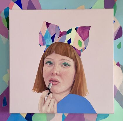 A beautiful red headed girl with bright green eyeshadow  and rosy cheeks is applying a peach colour lipstick. She is wearing an amazingly colourful and quirky headband with a big bow. She is dressed in colbolt blue and is posed against a lilac background.