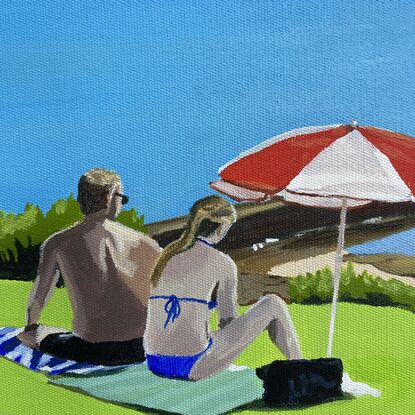 A man and woman wearing swimmers are sitting on towels on green grass under a red and white beach umbrella. An ocean pool and the greater ocean is in the background under a clear blue sky.  There are plants and shrubs on the left side with small bright orange flowers.