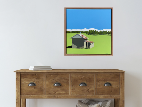 A small shed with a rusted roof casting a dark shadow to the left on a large bright green grass field, with distant trees. The sky is bright blue and there is a ribbon of white fluffy clouds on the horizon.