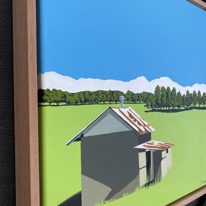 A small shed with a rusted roof casting a dark shadow to the left on a large bright green grass field, with distant trees. The sky is bright blue and there is a ribbon of white fluffy clouds on the horizon.