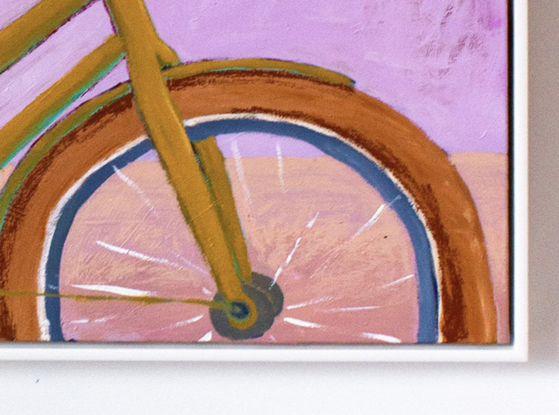 A crop of a woman riding a bike, wearing navy shorts, white shoes, pink socks and a blue and white striped t'shirt. Her shoulders and head are cropped out of the artwork. The vintage looking bike is a chartreuse colour with ochre tires, the background is a classic pink, the ground a pale peach. The artwork sits in a white floating frame.