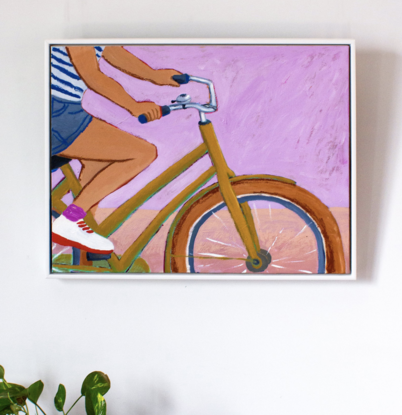 A crop of a woman riding a bike, wearing navy shorts, white shoes, pink socks and a blue and white striped t'shirt. Her shoulders and head are cropped out of the artwork. The vintage looking bike is a chartreuse colour with ochre tires, the background is a classic pink, the ground a pale peach. The artwork sits in a white floating frame.