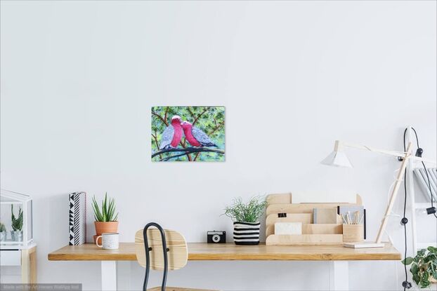 Two pink and grey galahs sitting on a branch in a loving and romantic stance. The background is various shades of green representing the leaves in the trees behind and blue sky filters through the leaves. The painting is framed in a light wooden floating frame.