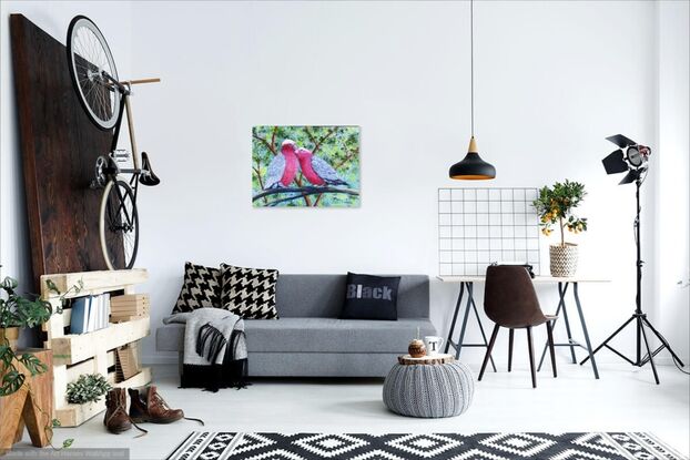 Two pink and grey galahs sitting on a branch in a loving and romantic stance. The background is various shades of green representing the leaves in the trees behind and blue sky filters through the leaves. The painting is framed in a light wooden floating frame.