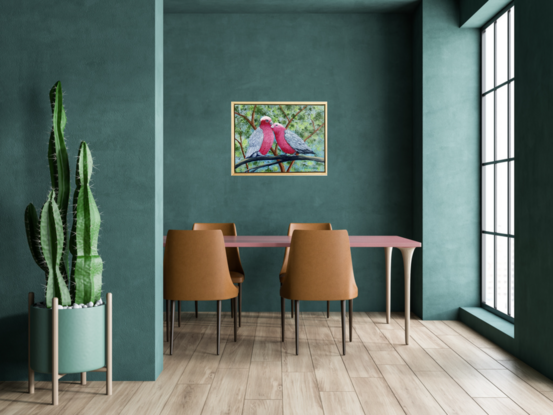 Two pink and grey galahs sitting on a branch in a loving and romantic stance. The background is various shades of green representing the leaves in the trees behind and blue sky filters through the leaves. The painting is framed in a light wooden floating frame.