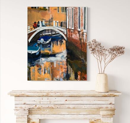 A couple of people sit with their knees on a step on a Venetian Bridge. There are a couple of boats in the water. And bright colours from the building behind them reflecting in the water.