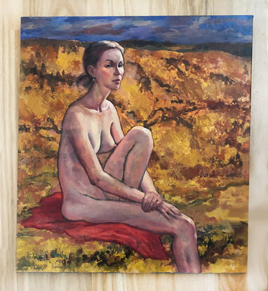 Oil painting of a female nude, in the countryside, sitting on a red cloth, looking at the viewer. The landscape is rough scrub and ochre terrain, in hues of brown, russet, orange and deep green. There is deep blue sky at the top of the painting. The painting is textured, and varnished.