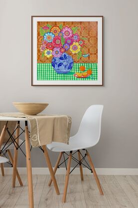 Colourful flowers in blue and white vase in front of red ochre patterned background on green checkered tablecloth with a pink plate of oranges.