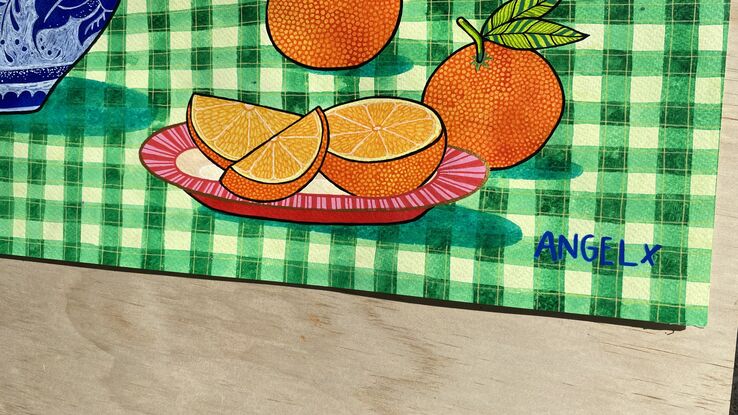 Colourful flowers in blue and white vase in front of red ochre patterned background on green checkered tablecloth with a pink plate of oranges.