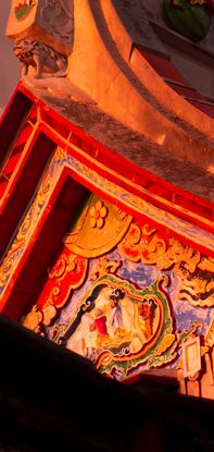 Swallow tail roof is drenched in golden sunlight, ornate features of figures and and floral patterns adorn the wall on the side of the house, beneath the roof. A grey storm cloud frames the roof and is lined with pale orange sunlight. dominating the top half of the picture is a deep blue sky.