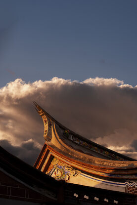 Swallow tail roof is drenched in golden sunlight, ornate features of figures and and floral patterns adorn the wall on the side of the house, beneath the roof. A grey storm cloud frames the roof and is lined with pale orange sunlight. dominating the top half of the picture is a deep blue sky.