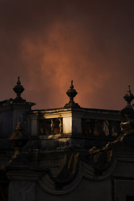 Red, hazy clouds hang above three minaret on the balcony of a western-style balcony. Most of the building is in shadow, except for once spot in the middle that has a circular highlight.