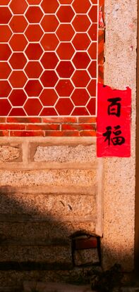 The front door of a house is surrounded by red banners with well wishes written on them. To the left of the image half of the tiled wall, beside the door, is in shadow which continues down into the courtyard at the front of the house. 

Above of the curve of the roof as a vibrant blue sky and the top of fluffy white cloud peeks above the roof tiles, inline with the door.