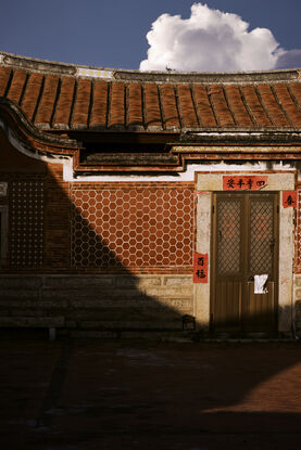 The front door of a house is surrounded by red banners with well wishes written on them. To the left of the image half of the tiled wall, beside the door, is in shadow which continues down into the courtyard at the front of the house. 

Above of the curve of the roof as a vibrant blue sky and the top of fluffy white cloud peeks above the roof tiles, inline with the door.