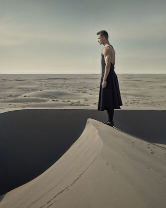 A lone man stands poised on the edge of a vast sand dune, his figure silhouetted against the muted tones of a grey sky. 