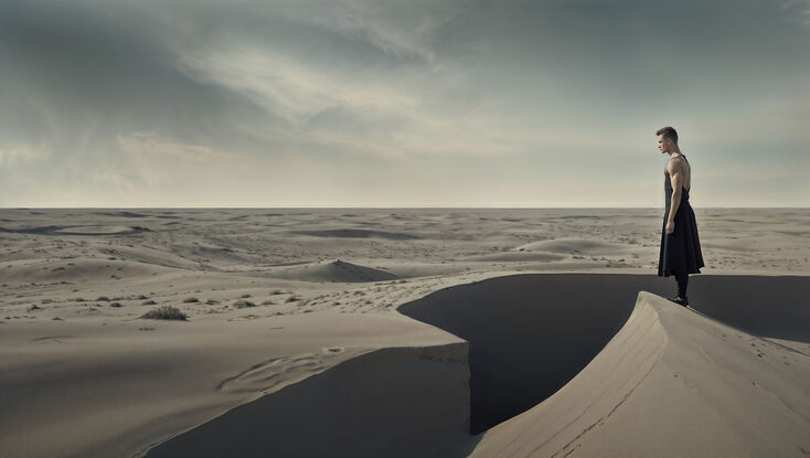 A lone man stands poised on the edge of a vast sand dune, his figure silhouetted against the muted tones of a grey sky. 