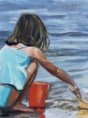 A girl in turquoise bathers squats on the edge of the ocean filling a red bucket with sand 