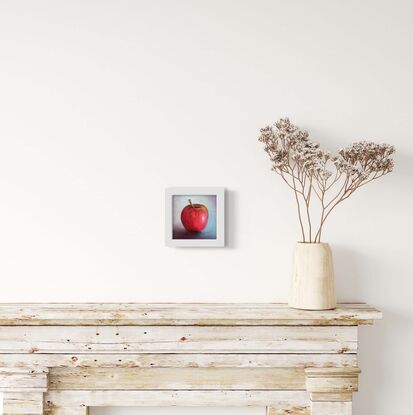 A small painting of a red apple with a blue background on a framed canvas.