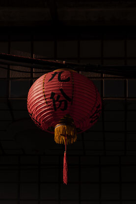 A red lantern is highlighted from the sunrise, it is set against a shadowy wall where many, small tiles can just be seen in the dark.