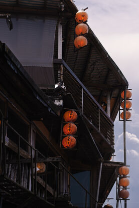 4 vertical rows of red lanterns adorn the side of a run down wooden building. Behind, the sky is filled with pink and blue hazy clouds.