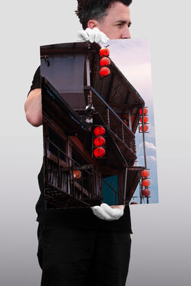 4 vertical rows of red lanterns adorn the side of a run down wooden building. Behind, the sky is filled with pink and blue hazy clouds.