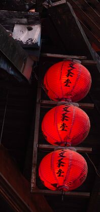 4 vertical rows of red lanterns adorn the side of a run down wooden building. Behind, the sky is filled with pink and blue hazy clouds.