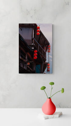 4 vertical rows of red lanterns adorn the side of a run down wooden building. Behind, the sky is filled with pink and blue hazy clouds.