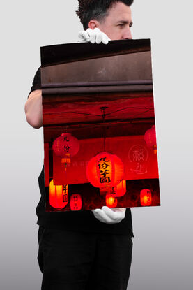 A collection of red, glowing lanterns hangs from the underside of a concrete ceiling. Above is the gritty face of the concrete building, intersected vertically with metal sheets.