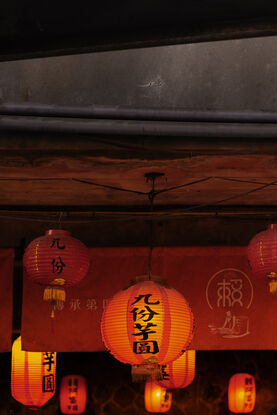 A collection of red, glowing lanterns hangs from the underside of a concrete ceiling. Above is the gritty face of the concrete building, intersected vertically with metal sheets.