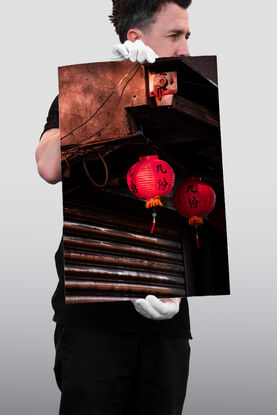 Two red lanterns hang in front of a steel roller door. Both gentle sway in the warm breeze. The front lantern casts a shadow over side of the back lantern, as it is bathed in light. Above the lanterns the weathered concrete of the house glows dark orange from the sun; wires cross it's face, old drill holes, metal rods and a shining steel casing adorn the surface.