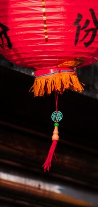 Two red lanterns hang in front of a steel roller door. Both gentle sway in the warm breeze. The front lantern casts a shadow over side of the back lantern, as it is bathed in light. Above the lanterns the weathered concrete of the house glows dark orange from the sun; wires cross it's face, old drill holes, metal rods and a shining steel casing adorn the surface.