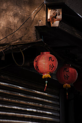 Two red lanterns hang in front of a steel roller door. Both gentle sway in the warm breeze. The front lantern casts a shadow over side of the back lantern, as it is bathed in light. Above the lanterns the weathered concrete of the house glows dark orange from the sun; wires cross it's face, old drill holes, metal rods and a shining steel casing adorn the surface.