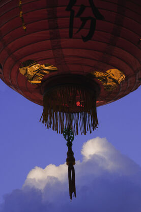 A dark, red lantern dominates the top half of the photograph, it floats in the air as sunlight catches the golden foil on it's underside. The tassle and skirt of the lantern bristle and sway in the breeze. In the dark blue sky behind it is a cloud that is reminiscent of a snow capped mountain - soft white light crowns it's peak.