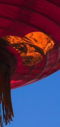 A dark, red lantern dominates the top half of the photograph, it floats in the air as sunlight catches the golden foil on it's underside. The tassle and skirt of the lantern bristle and sway in the breeze. In the dark blue sky behind it is a cloud that is reminiscent of a snow capped mountain - soft white light crowns it's peak.