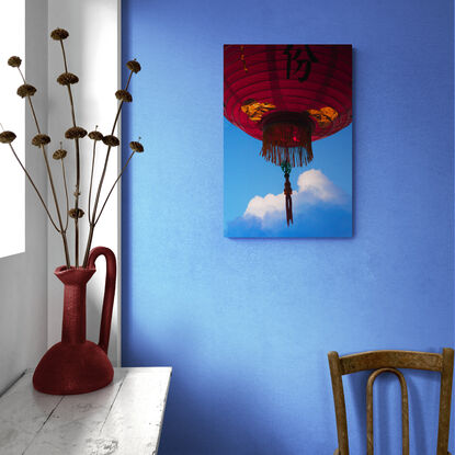 A dark, red lantern dominates the top half of the photograph, it floats in the air as sunlight catches the golden foil on it's underside. The tassle and skirt of the lantern bristle and sway in the breeze. In the dark blue sky behind it is a cloud that is reminiscent of a snow capped mountain - soft white light crowns it's peak.