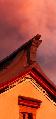 Pink, cotton candy rain clouds float above the corner of a house. The wall of the house is made orange by the setting sun as the corner of the roof reaches up towards the sky like a mountain peak.