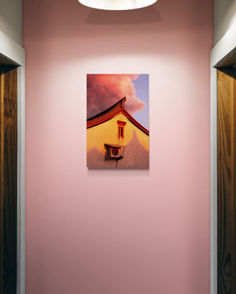 Pink, cotton candy rain clouds float above the corner of a house. The wall of the house is made orange by the setting sun as the corner of the roof reaches up towards the sky like a mountain peak.