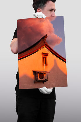 Pink, cotton candy rain clouds float above the corner of a house. The wall of the house is made orange by the setting sun as the corner of the roof reaches up towards the sky like a mountain peak.