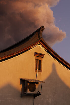 Pink, cotton candy rain clouds float above the corner of a house. The wall of the house is made orange by the setting sun as the corner of the roof reaches up towards the sky like a mountain peak.