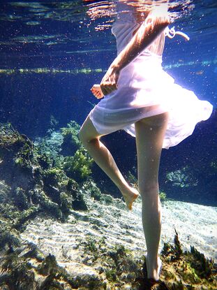 Young woman spinning underwater in white dress whilst standing on the sandy floor.